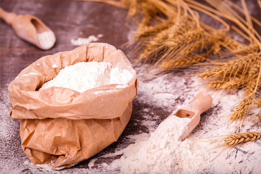 Mąka pełnoziarnista w papierowej torebce i drewnianej łyżce, z kłosami zboża na tle drewnianego stołu﻿